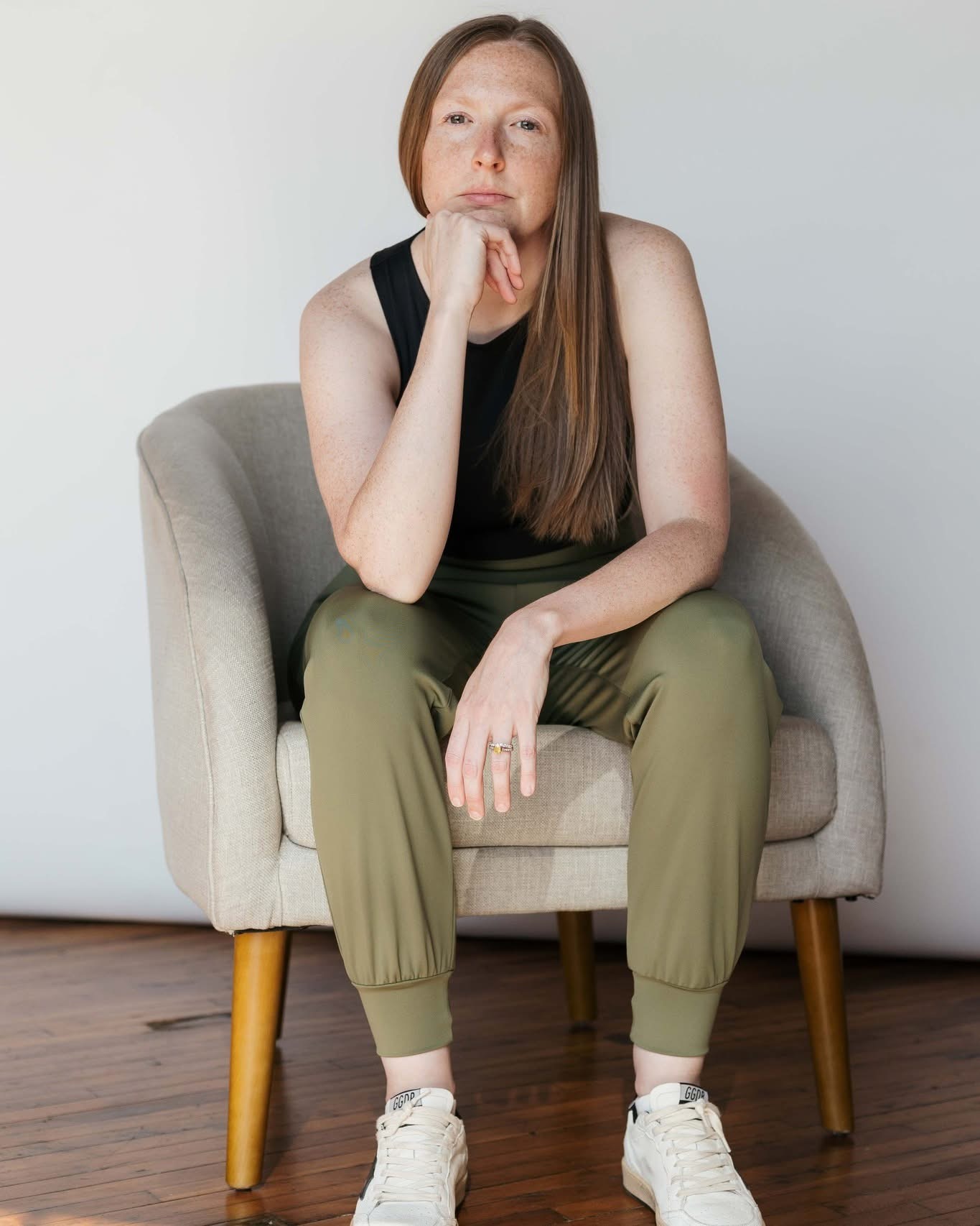 Leslye Young seated in a linen armchair on a wood floor, wearing olive joggers and a black tank, chin resting on her hand.