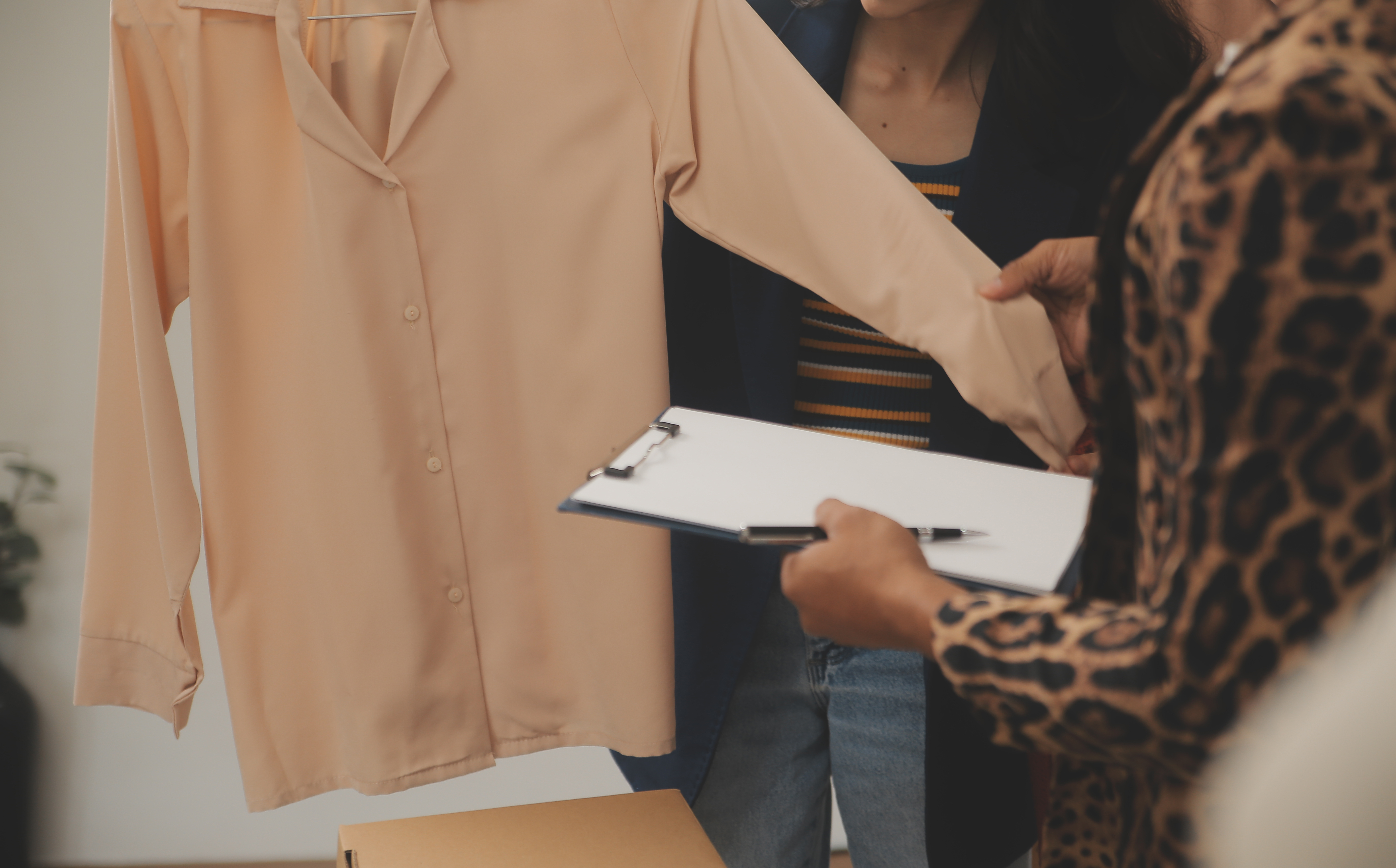 Two people reviewing a peach button-down sample on a hanger, clipboard in hand for written feedback.