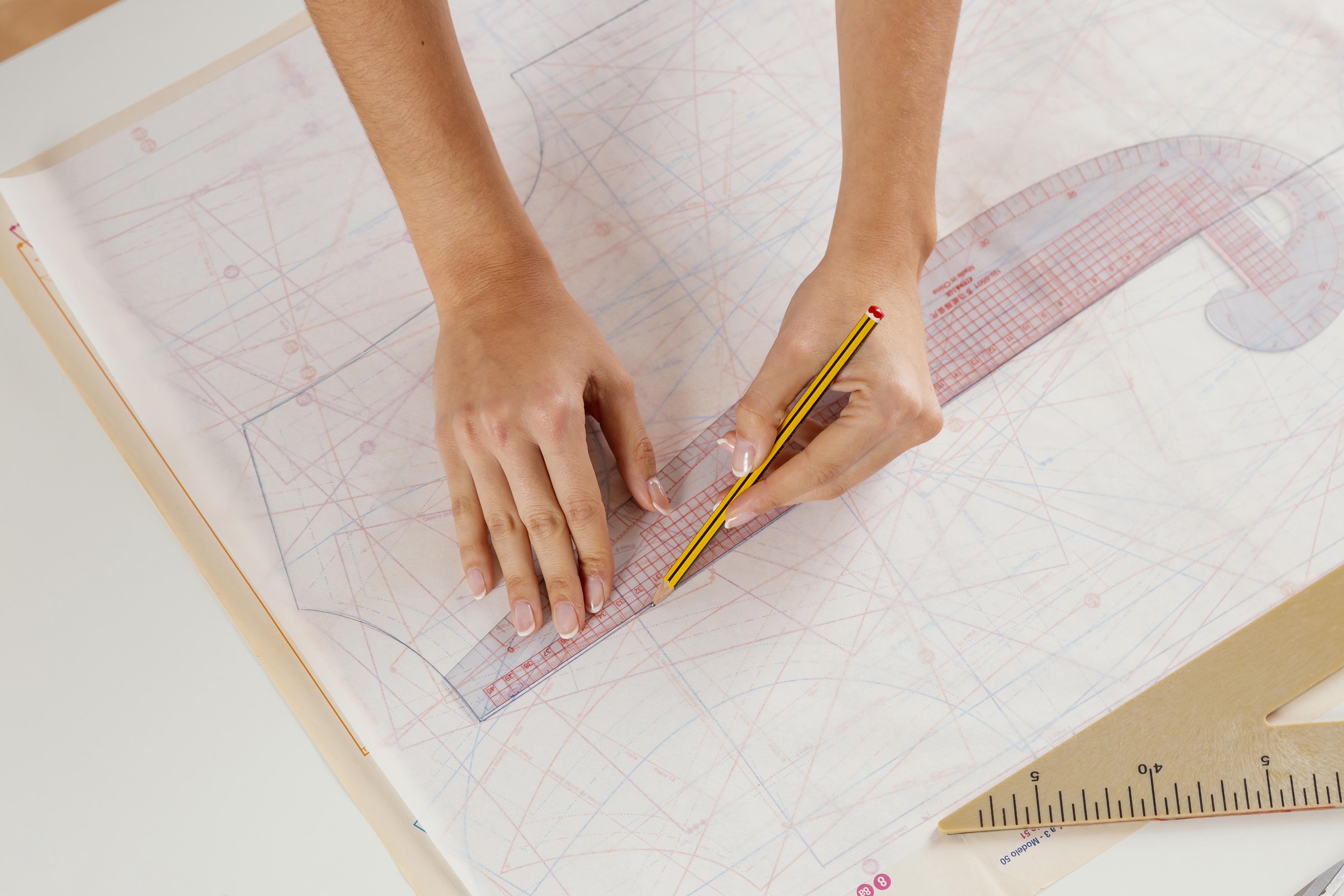 Hands drafting a garment pattern with a clear ruler and pencil on overlaid paper patterns.