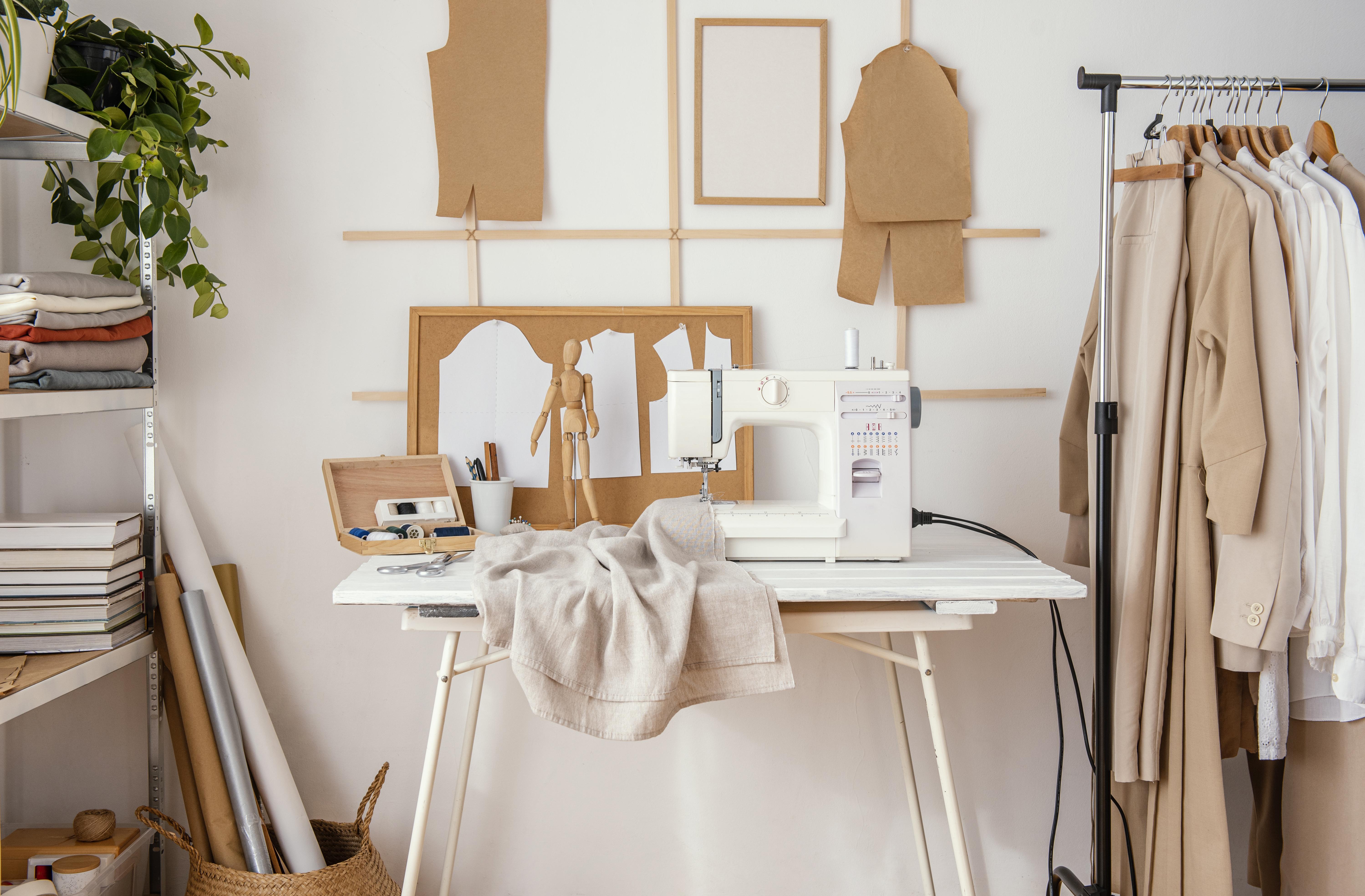 Tailoring studio with sewing machine, pattern pieces pinned to a corkboard, and finished neutral-toned garments on a rack.